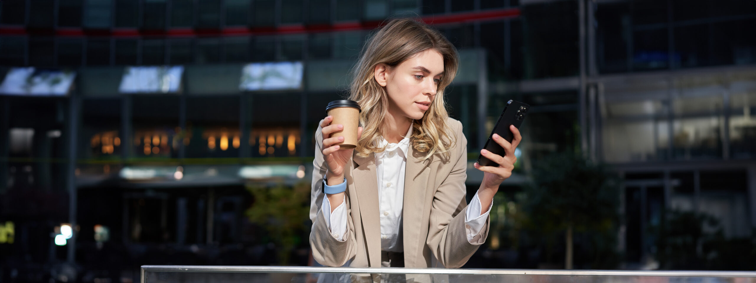 A young businesswoman in a blazer stands outside a modern office building, holding a coffee cup and smartphone, focused on online shopping.