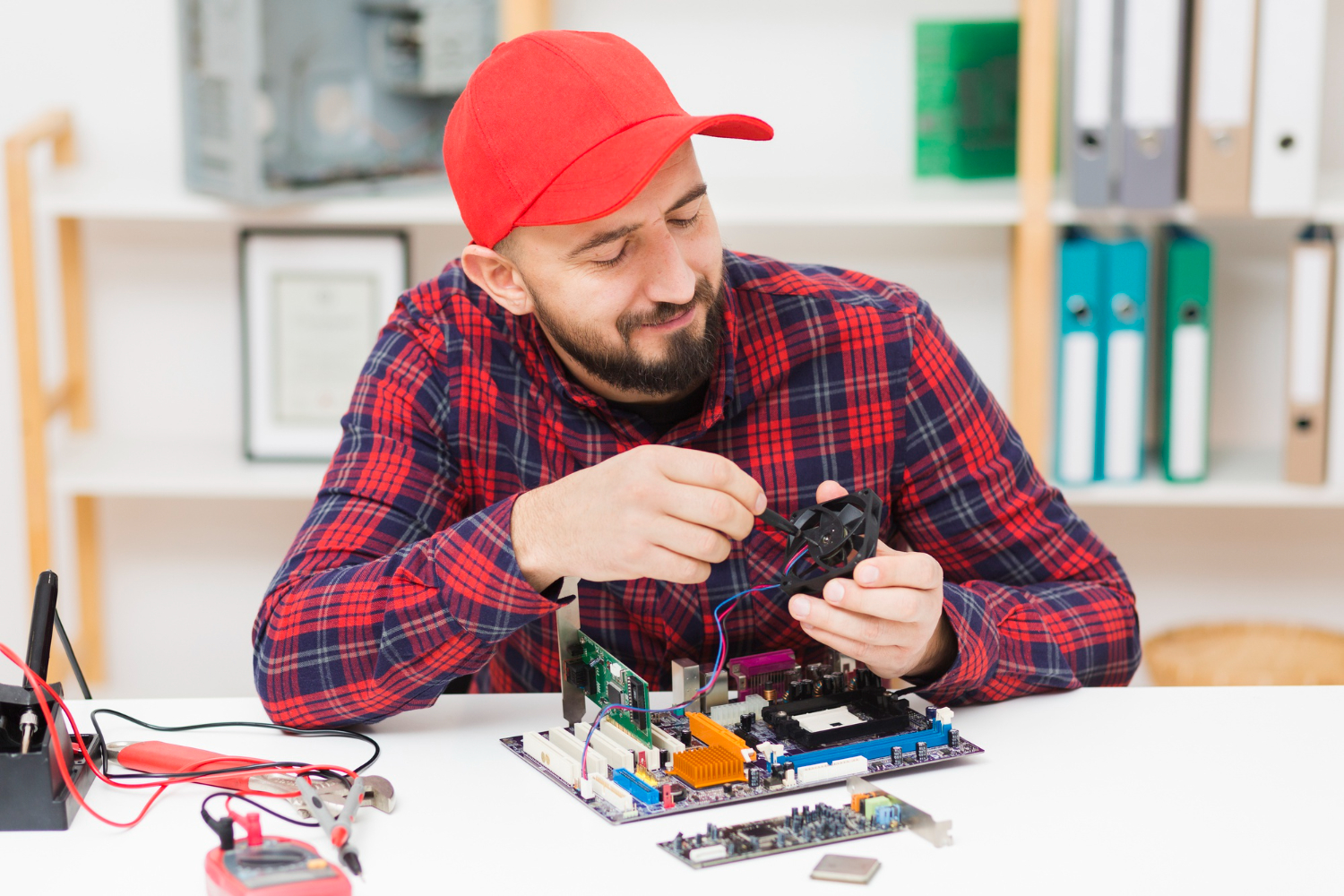 A man in a red cap and plaid shirt is assembling or repairing a computer motherboard, providing hardware solutions. The background shows an office with shelves of binders.